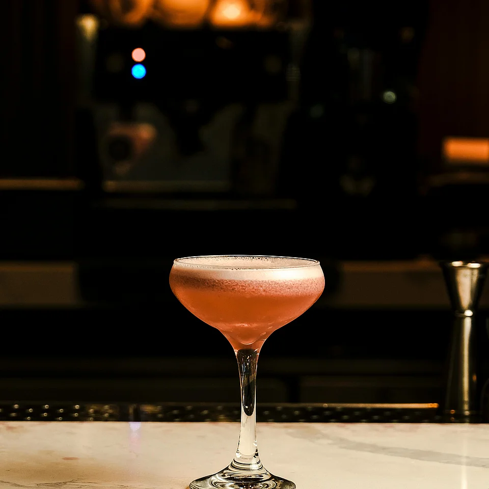 Ein Glas mit pinkfarbenem Cocktail steht auf einer hellen Marmoroberfläche vor dunklem Hintergrund in einer Bar.