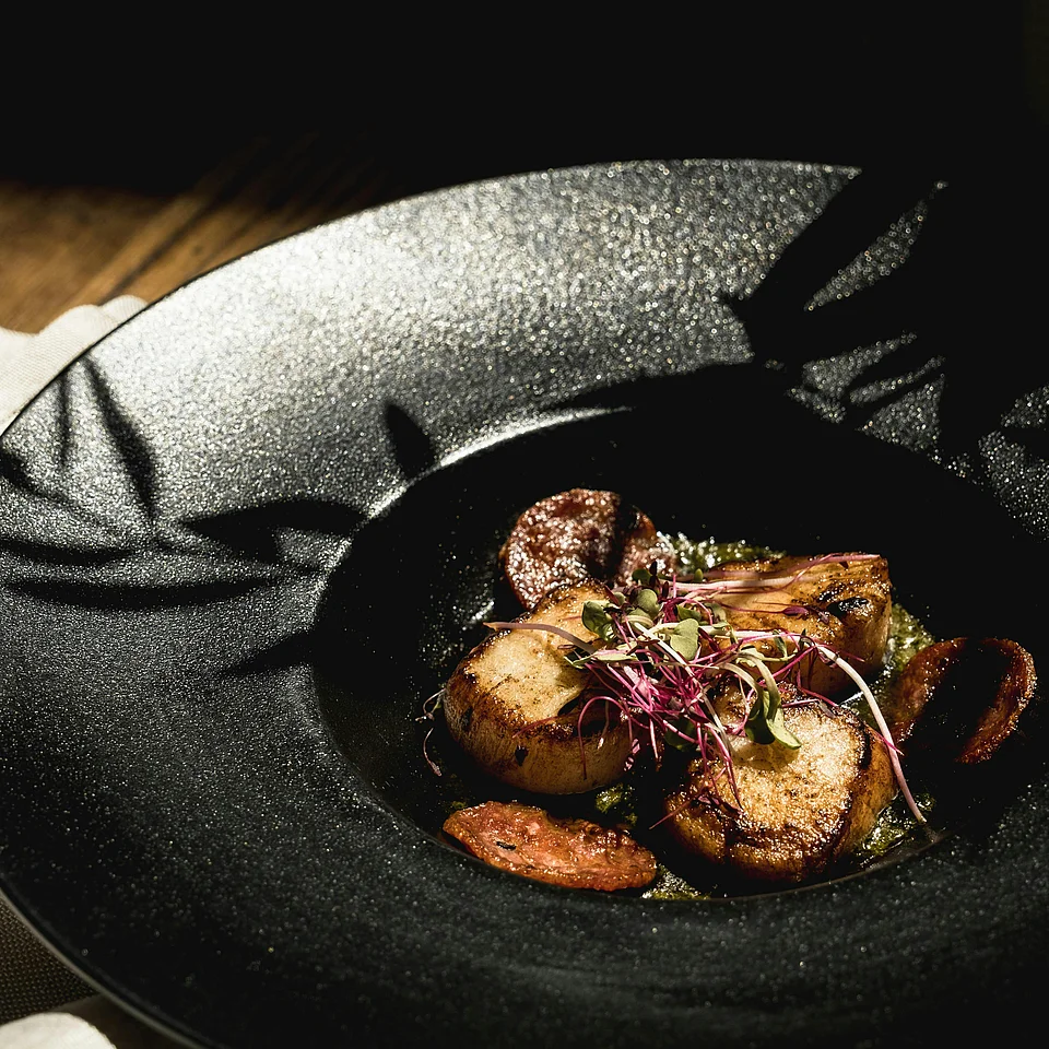 Elegante schwarze Schale mit gebratenen Kartoffeln, Möhrenscheiben und frischen Kräutern dekoriert, auf einem dunklen Holztisch.