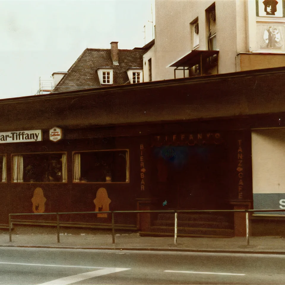 Fassade einer dunklen Tanzbar namens Tanz-Bar-Tiffany entlang einer Straßenecke mit einem kleinen Schild für Schlecker nebenan.
