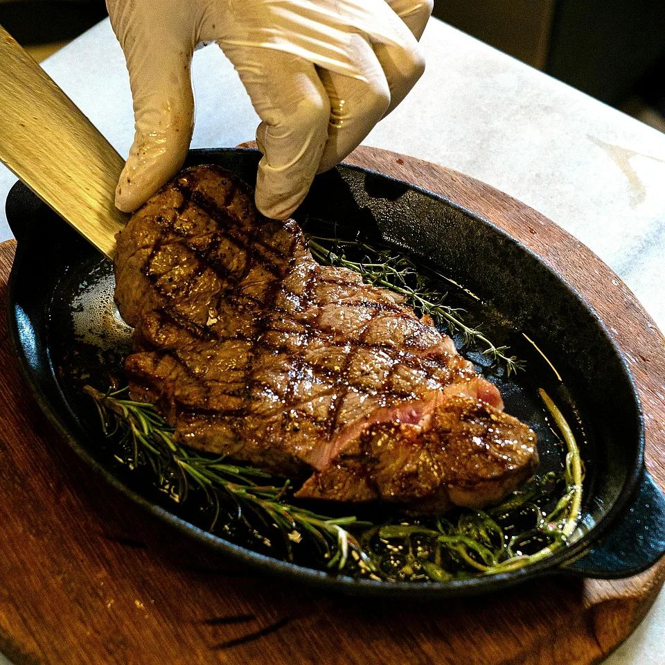 Hand in Einweghandschuh hebt ein gegrilltes Steak aus einer Pfanne mit Kräutern auf einem Holzbrett