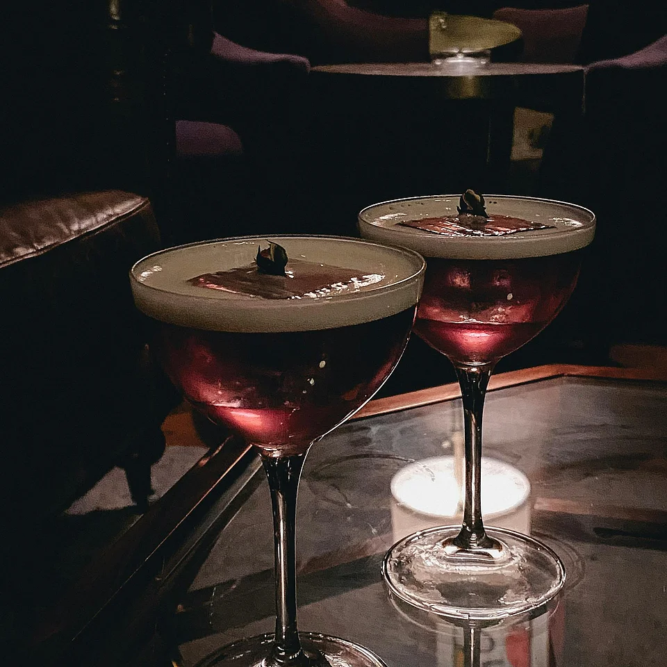 Zwei Cocktails in eleganten Gläsern auf einem Glastisch in einer gedämpft beleuchteten Bar mit Ledermöbeln im Hintergrund.