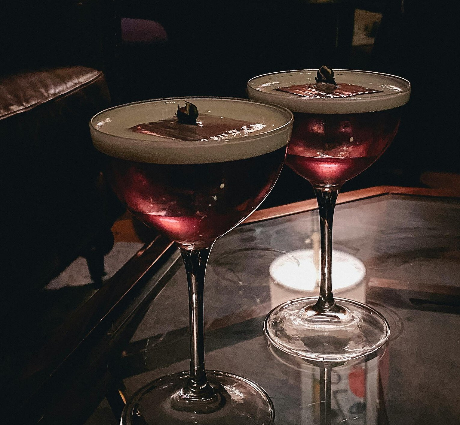 Zwei Cocktails in eleganten Gläsern auf einem Glastisch in einer gedämpft beleuchteten Bar mit Ledermöbeln im Hintergrund.