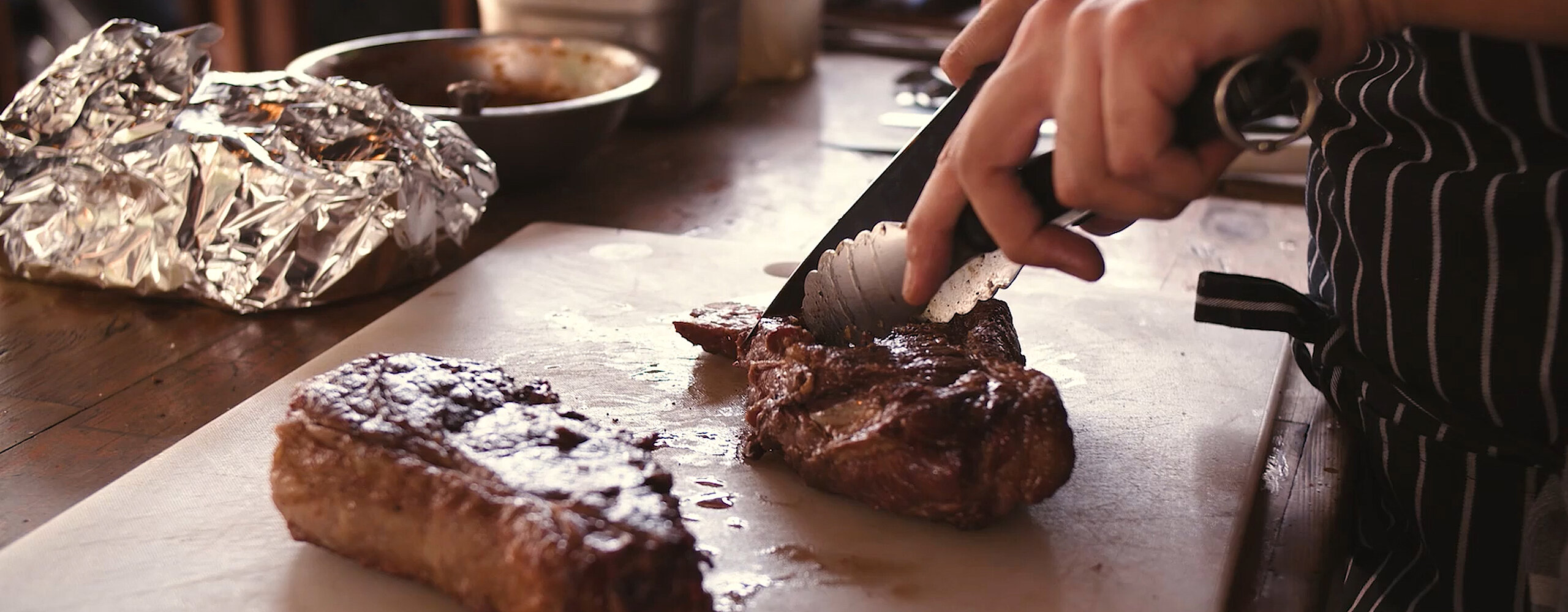 Eine Hand schneidet ein Stück gegrilltes Fleisch auf einem Holzbrett, daneben liegt ein weiteres Stück Fleisch.
