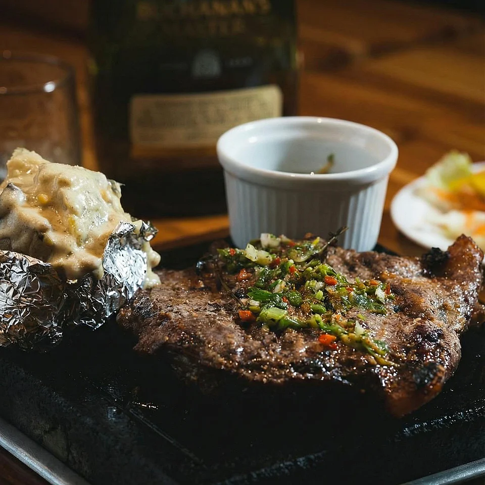 Gegrilltes Steak mit Kräuterbutter, gebackene Kartoffel mit saurer Sahne und ein kleines Gefäß mit Sauce auf einem schwarzen Serviertablett, im Hintergrund eine Flasche Whiskey.