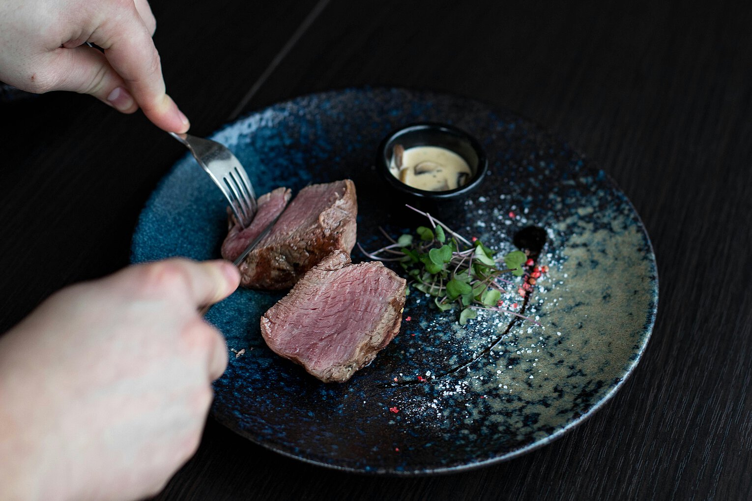 Hände schneiden ein Stück medium gebratenes Steak auf einem dunklen, dekorativen Teller mit einer kleinen Portion Sauce und etwas Mikrogrün.