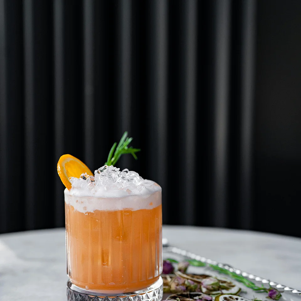 Ein mit Crushed Ice gefüllter Cocktail in einem facettierten Glas, garniert mit einer Orangenscheibe und einem Rosmarinzweig, auf einem Marmoruntergrund mit getrockneten Blüten daneben.