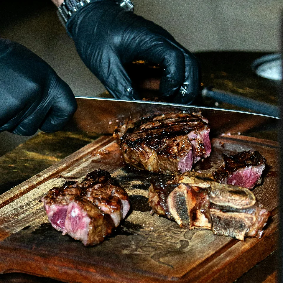Hände mit schwarzen Handschuhen schneiden ein Stück gegrilltes Steak auf einem Holzbrett.