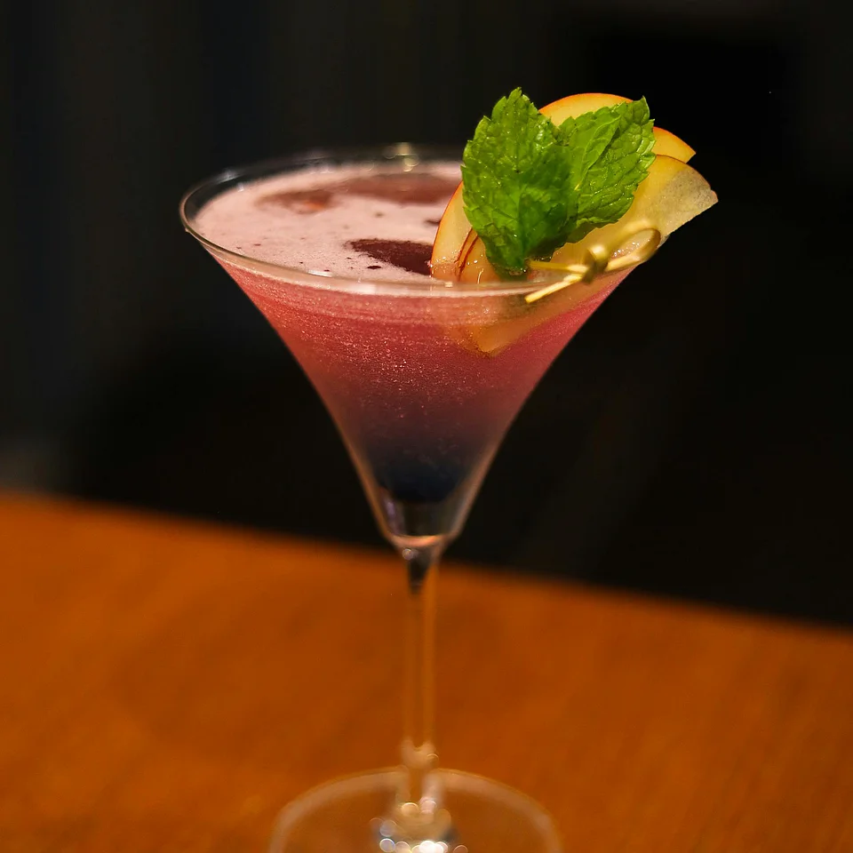Cocktail in einem Martiniglas mit rosa Farbe, garniert mit Minzblättern und Fruchtscheiben auf einem Holztisch.